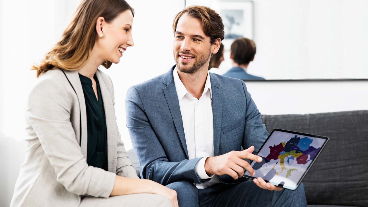 Consilier financiar discutând cu o clientă și prezentând informații pe o tabletă într-un cadru profesional.